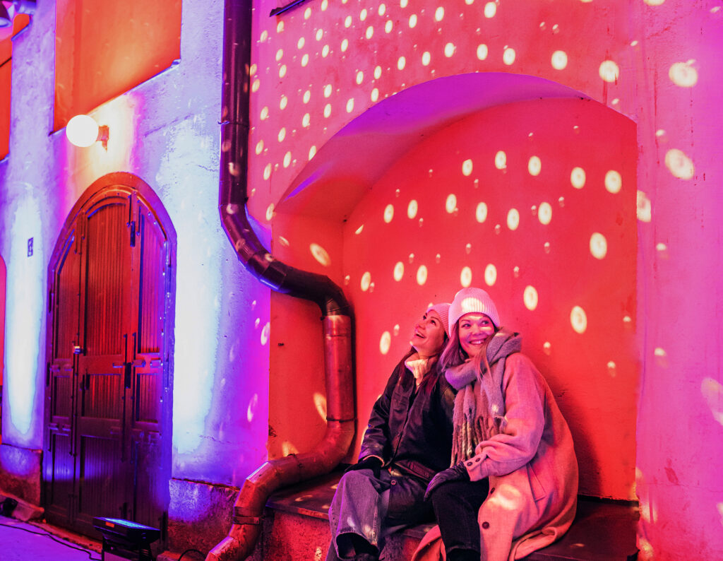 Kaksi ihmistä istuskelee värivaloin koristetulla Finlaysonin alueella talviaikaan.